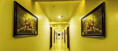 A hotel corridor with yellow walls, framed artwork on the walls, and a long hallway leading to a door at the end.. Hotel Amar Vilas & Resort Bharatpur