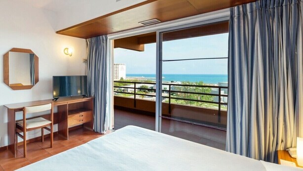 Un dormitorio con una cama grande, un escritorio, una silla, un televisor y un balcón con vista al mar.