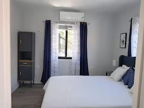 Ein Schlafzimmer mit Bett, Schrank, einem Fenster mit Vorhängen und einer Klimaanlage.