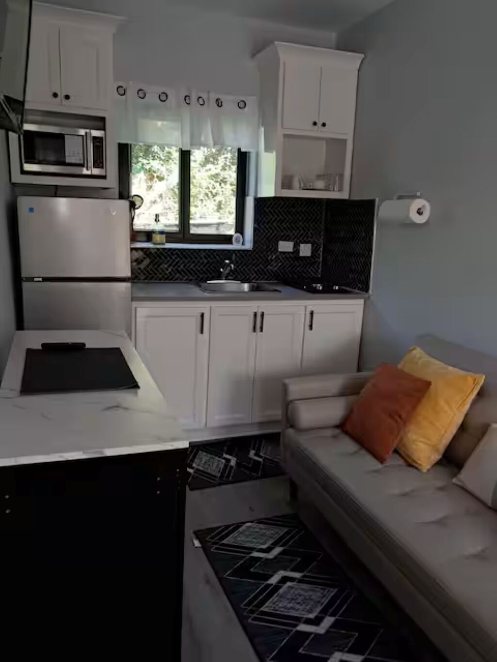 Una cocina compacta con armarios blancos, microondas y refrigerador. Se ve un ventanal con vistas a la vegetación.