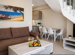 A modern living room with a brown sofa, a dining area with white chairs, a kitchen in the background, and a painting of a beach scene. - Casale Luxury House III - Secret Garden Maisonette (Sithonia)
