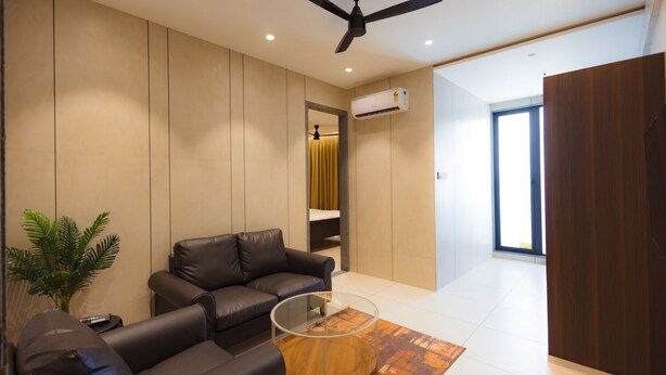 A modern living room with a sofa, a glass coffee table, a ceiling fan, and an air conditioning unit. - Nirrtul Crystal 1BHK Apartment Cozy & Quite (Surat)