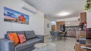 Un salon moderne avec un canapé gris, deux petites tables d’appoint rondes, un coin repas avec des chaises, une cuisine avec des meubles en bois et un climatiseur fixé au mur.