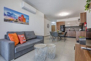 Ein modernes Wohnzimmer mit grauem Sofa, zwei runden Beistelltischen, einer Essecke mit Stühlen, einer Küche mit Holzschränken und einer wandmontierten Klimaanlage.