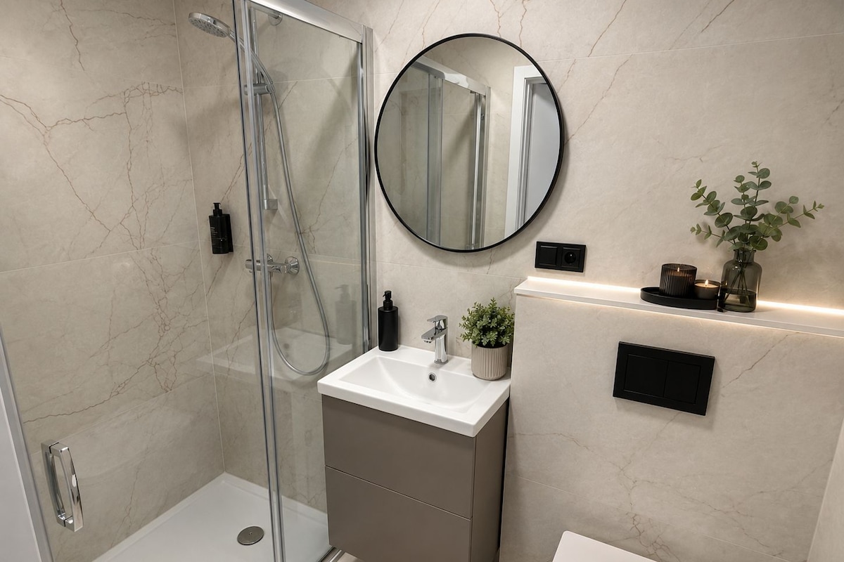 Un baño moderno con ducha envuelta en vidrio, lavamanos blanco y espejo redondo.