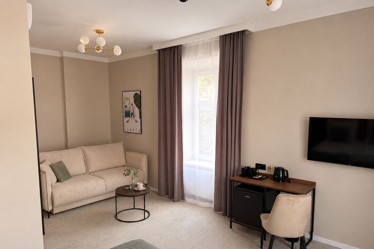Habitación de hotel con sofá, una pequeña mesa redonda con una planta, escritorio con silla, televisor y ventana con cortinas.