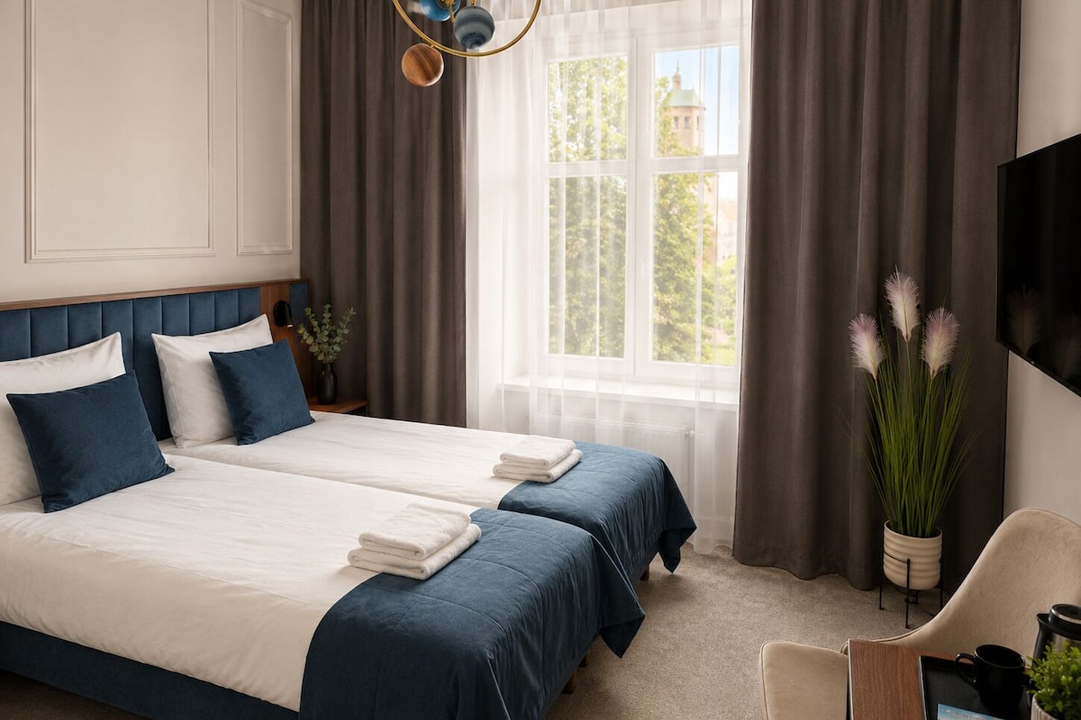 A hotel room with two beds, a large window with curtains, a flat-screen TV, a desk with a chair, and a potted plant.
