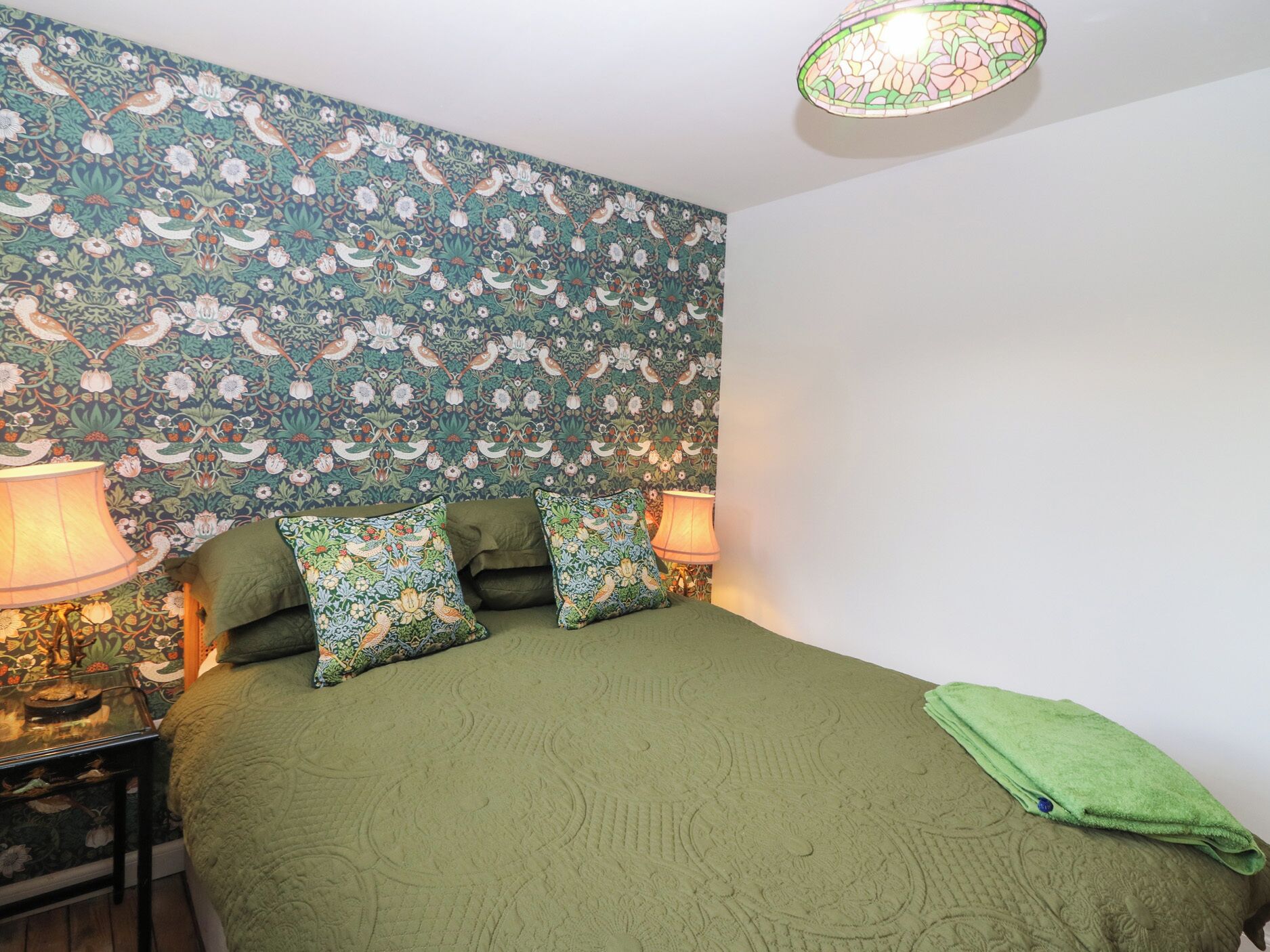 A bedroom with a floral wallpaper, green bedding, two bedside lamps, and a green towel on the bed.