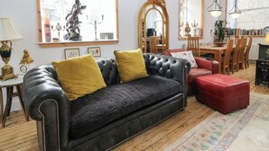 Una sala de estar con un sofá de cuero negro, un reposapiés rojo, una mesa de comedor con sillas, una araña y un espejo grande.