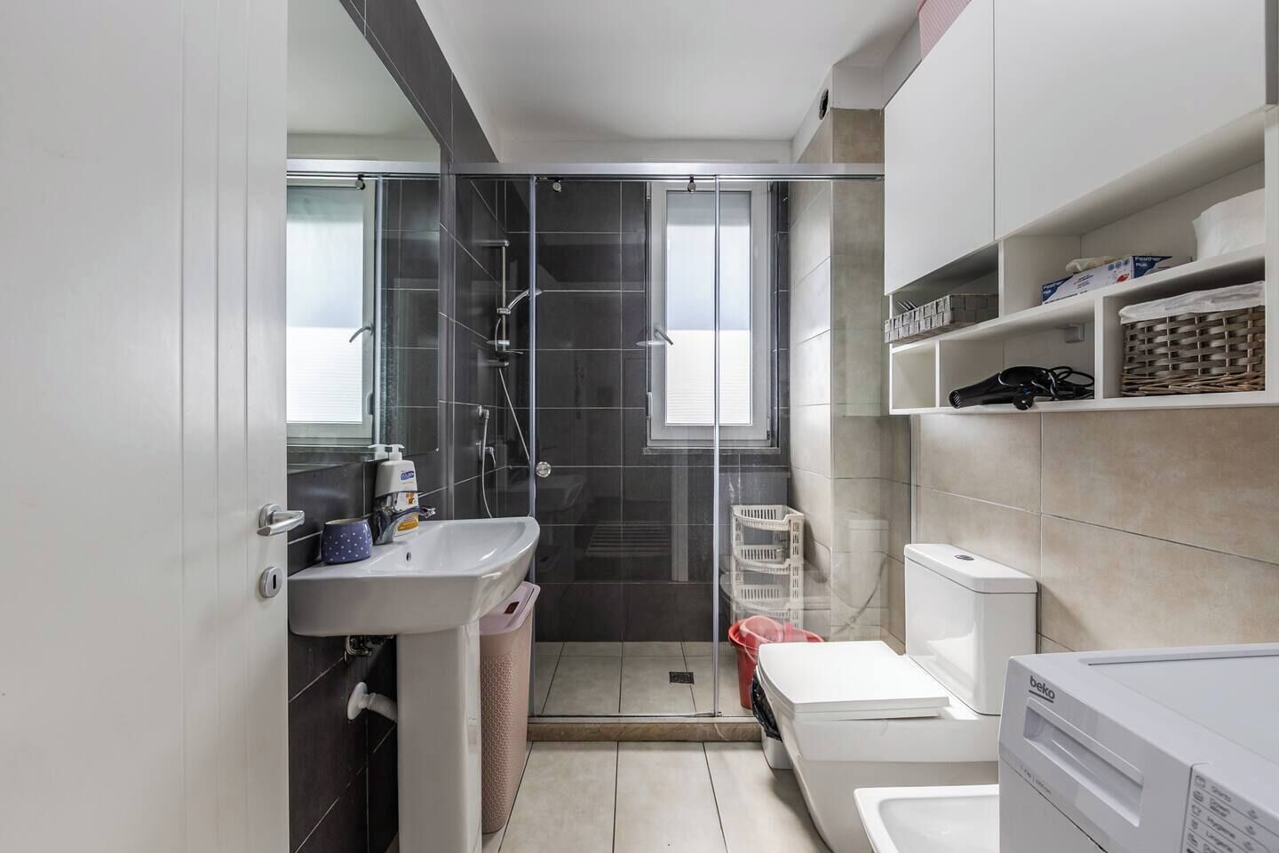 A modern bathroom with a glass shower enclosure, a sink with a soap dispenser, a toilet, and a washing machine.