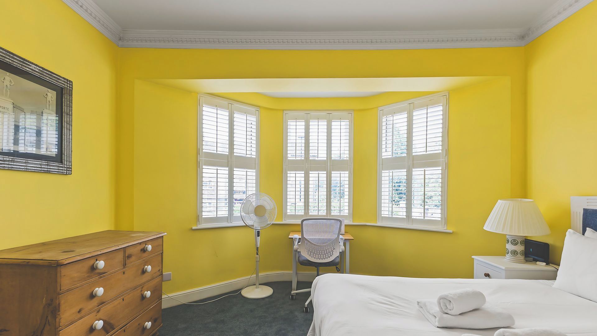 A hotel room with a corner window, a wooden dresser, a desk with a lamp, and a bed with a rolled towel.