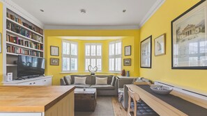 A living room with a yellow wall, a bookshelf, a television, a sofa, a coffee table, and a dining area with a table and chairs. - Fantastic 1BDR Flat in Chiswick w Spacious Garden (Greater London)
