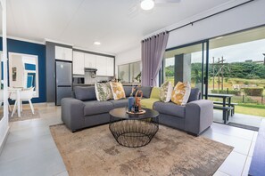 A modern living room with a grey sofa, a round coffee table, and a view of a garden through large windows. - Ballito Village Estate (Ballito)