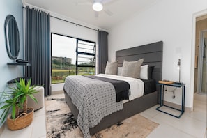 A modern bedroom with a large bed, a television, a side table, and a view of the outdoors. - Ballito Village Estate (Ballito)