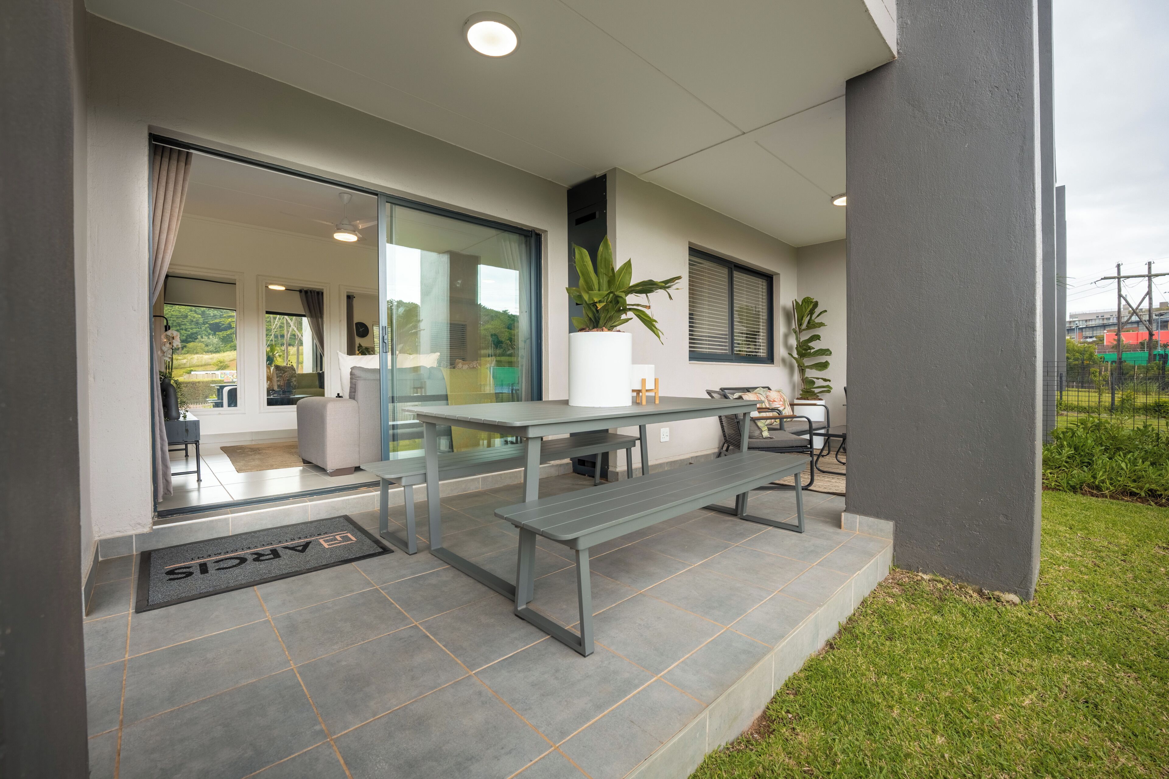 A modern outdoor patio with a table and benches, a potted plant, and a glass door leading to an interior space.