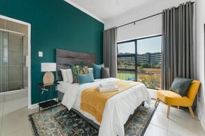 A modern bedroom with a large bed, a view of the city, and a balcony. - Ballito Village Estate (Ballito)