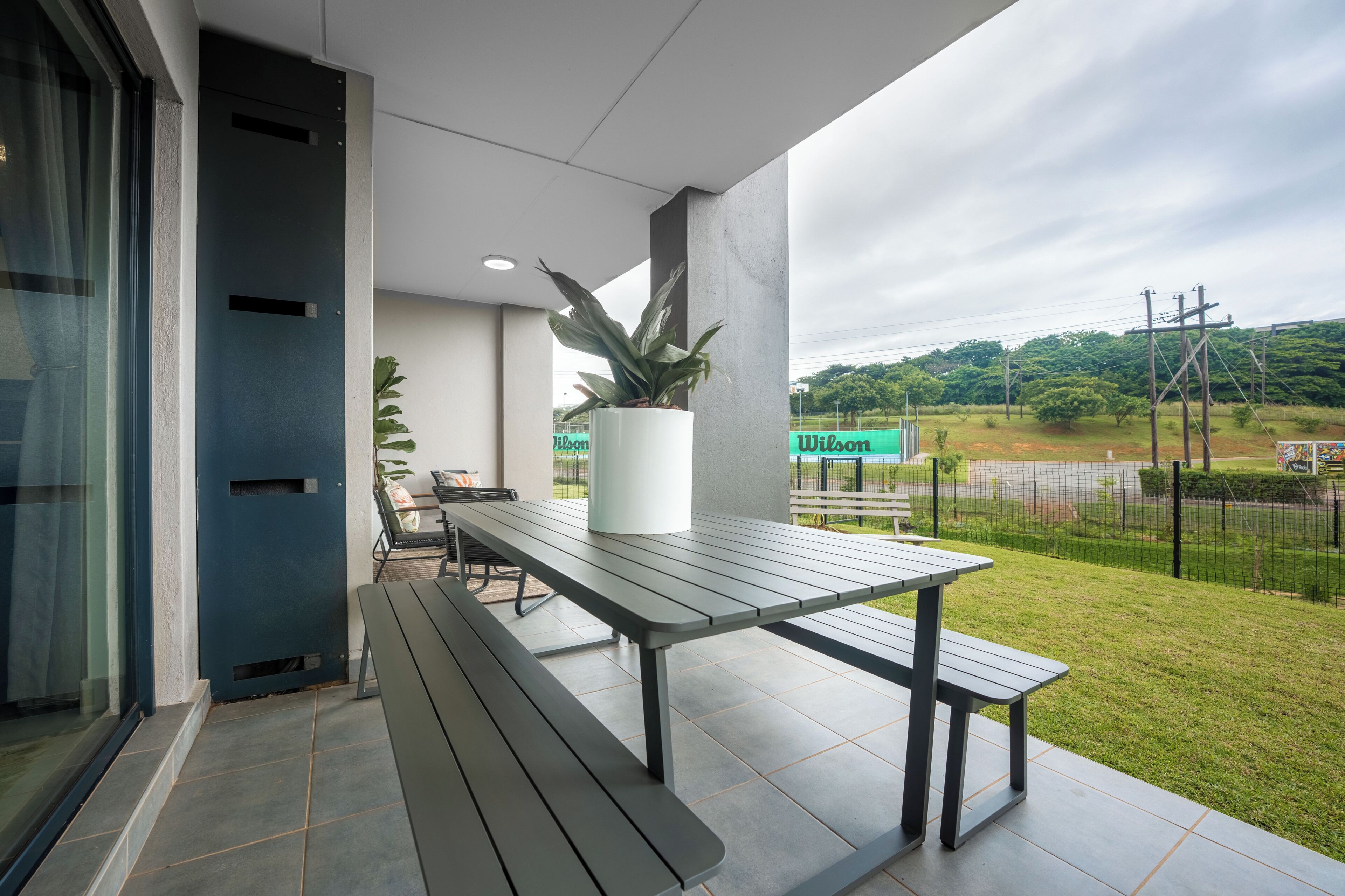 Een terras buiten met een tafel, bankjes, een plant in pot en uitzicht op een groen gebied met een hek.