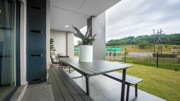 Een terras buiten met een tafel, bankjes, een plant in pot en uitzicht op een groen gebied met een hek.