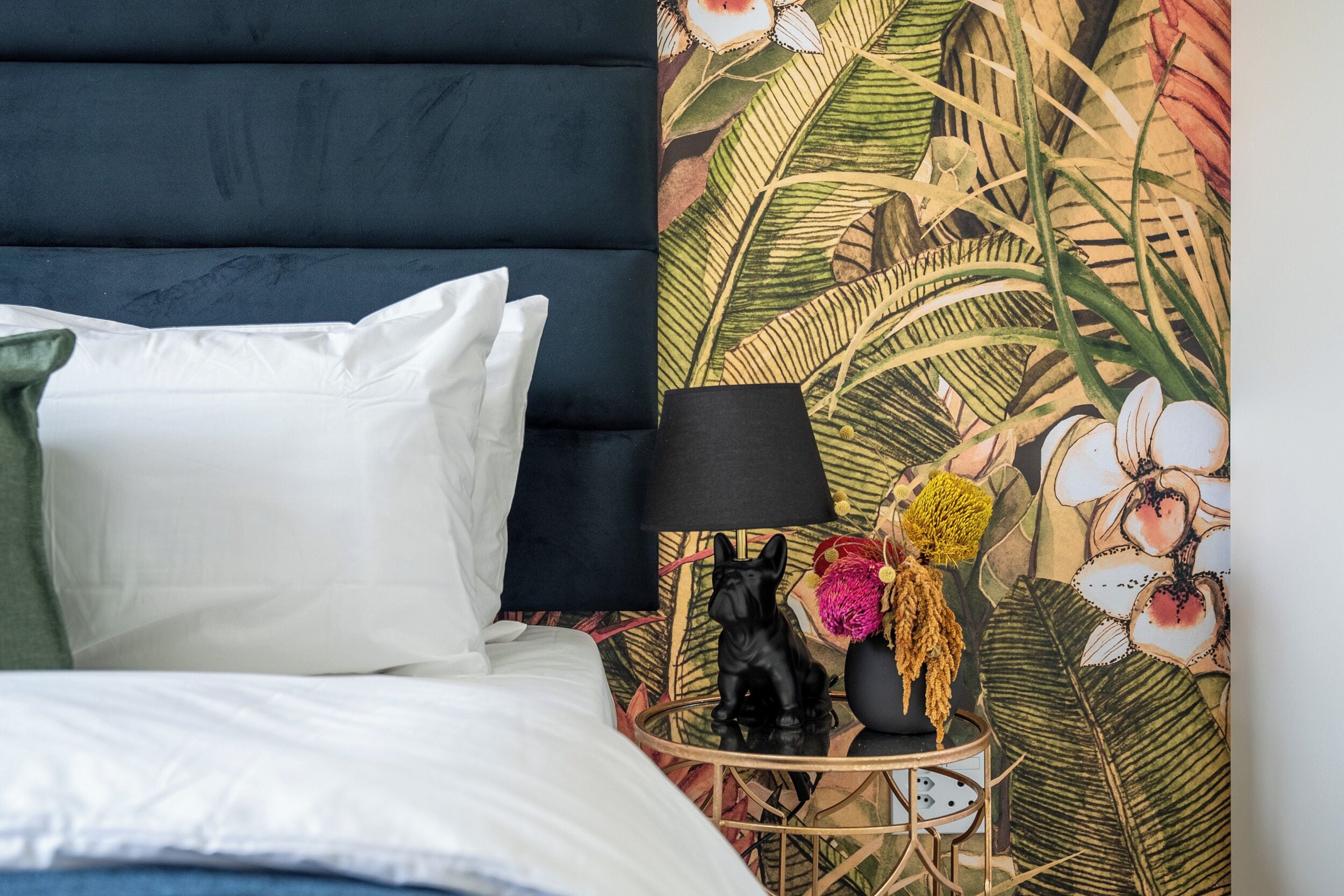 Ein Bett mit weißer Bettwäsche, ein dunkles Kopfteil und ein Nachttisch mit einer Lampe, einer kleinen schwarzen Figur und einer Vase mit Blumen.