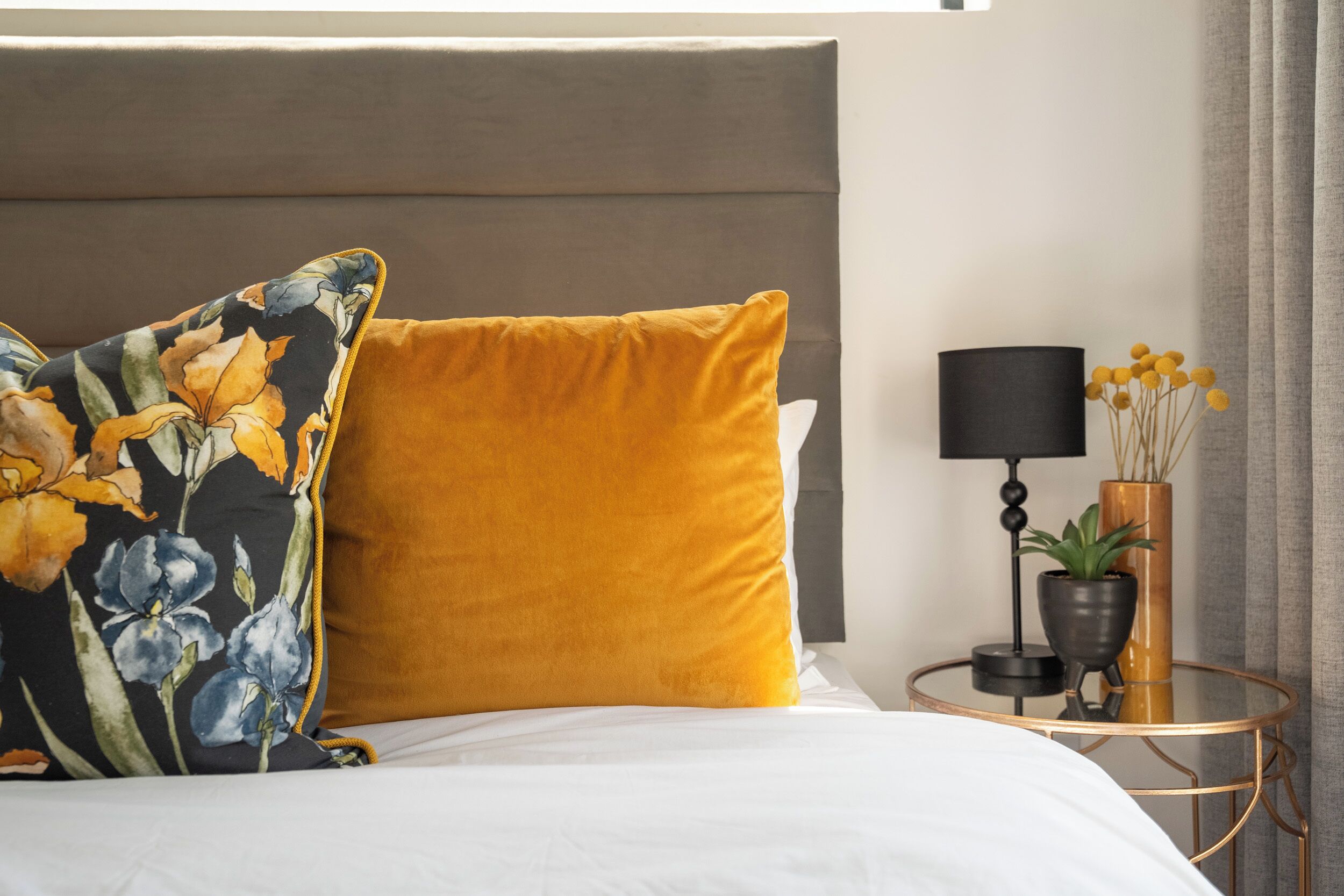 Una cama con un almohadón floral, un almohadón color mostaza, una mesita redonda con una lámpara y una planta.
