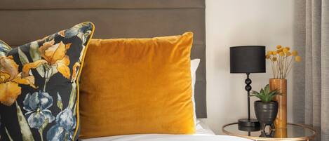 Una cama con un almohadón floral, un almohadón color mostaza, una mesita redonda con una lámpara y una planta.