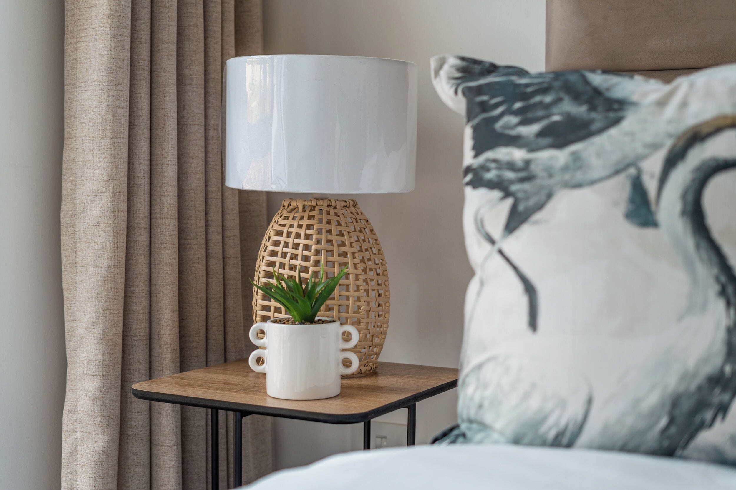 A bedside table with a lamp, a small potted plant, and a pillow with a bird design.