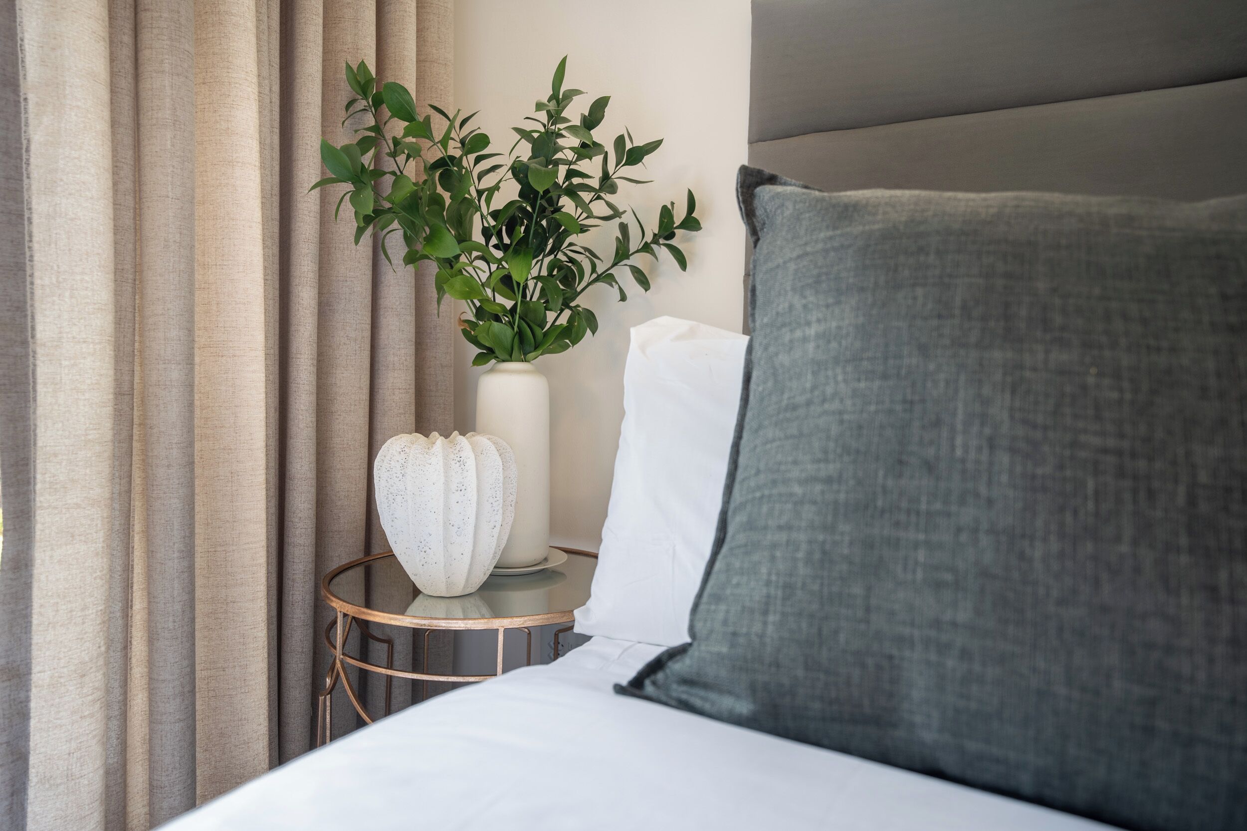 Een netjes opgemaakt bed met een grijs kussen, een witte vaas met een plant, en een rond nachtkastje met een wit object erop.