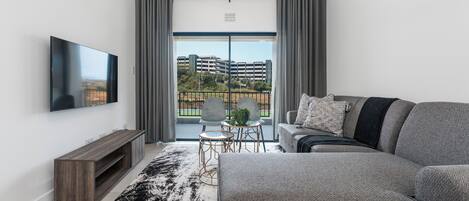 Un salon moderne avec un téléviseur à écran plat, un canapé gris et une vue sur un immeuble par une grande fenêtre.