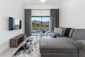 A modern living room with a flat-screen TV, grey sofa, and a view of a building through a large window. - Ballito Village Estate (Ballito)
