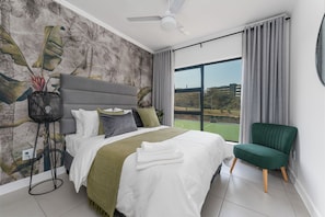 A modern hotel room with a large bed, a green armchair, a ceiling fan, and a view of the outdoors. - Ballito Village Estate (Ballito)