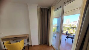 A balcony with a chair and a table, a sliding glass door, and a view of buildings and greenery. - El Rincon 11 - One Bed (Los Cristianos)
