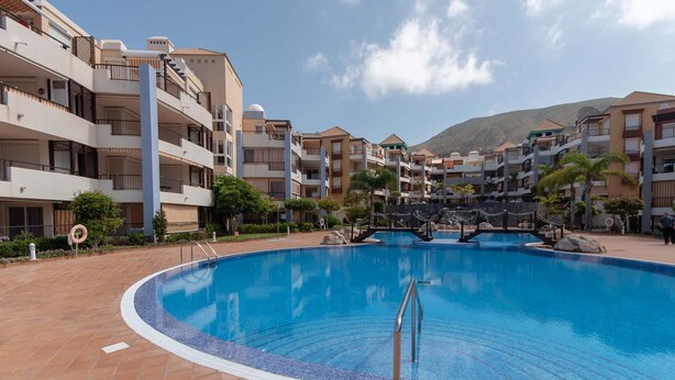 A swimming pool surrounded by multi-story buildings with balconies, a bridge over the pool, and a mountainous backdrop. - El Rincon 11 - One Bed (Los Cristianos)