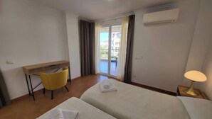 A hotel room with two beds, a desk, a chair, a lamp, and a view of the outside. - El Rincon 11 - One Bed (Los Cristianos)