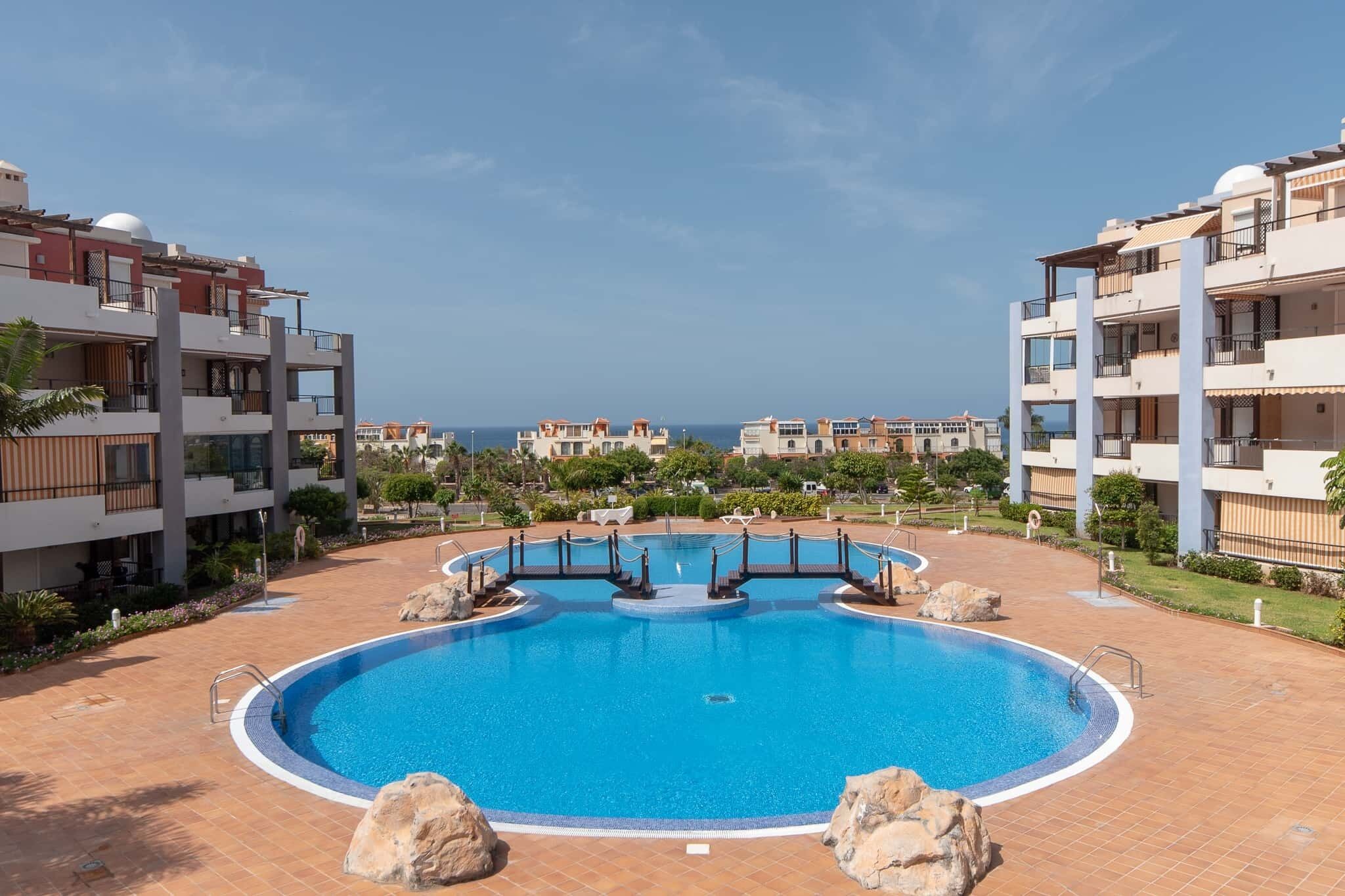 A swimming pool surrounded by modern buildings with balconies, a clear sky, and a distant view of the sea.