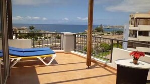 A balcony with a blue lounge chair, a wicker table with a red flower, and a view of the sea and buildings. - El Rincon 11 - One Bed (Los Cristianos)
