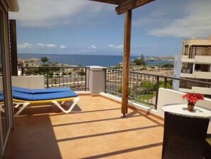 Ein Balkon mit blauem Liegestuhl, einem Korbstuhl mit einer roten Blume und Blick auf das Meer und die Gebäude.