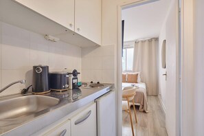 Eine kleine Küche mit Spüle, Herd und Kaffeemaschine. Im angrenzenden Raum sind ein Bett und ein Sessel zu sehen.
