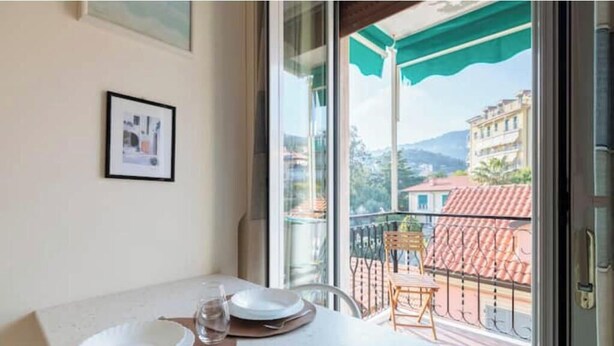 En balkon med et bord dækket til to personer, et billede i ramme på væggen og udsigt til bygninger og bakker.