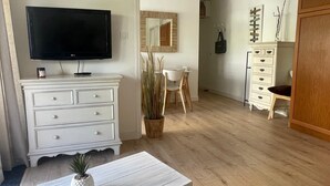 Una habitación con televisión de pantalla plana, cómoda blanca, aparador de madera, mesa de comedor con sillas y una planta en maceta.