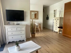 Ein Zimmer mit einem Flachbildfernseher, einer weißen Kommode, einem Holzschrank, einem Esstisch mit Stühlen und einer Topfpflanze.