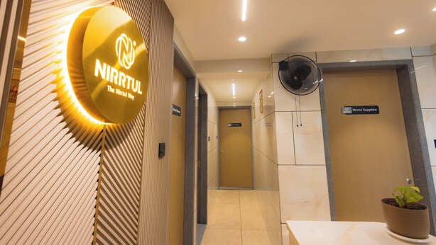 A hotel reception area with a modern design, featuring a reception desk, a potted plant, a wall-mounted fan, and a sign indicating the hotel's name. - Nirrtul Sapphire Studio Apartment Specious Cozy & Quite (Surat)