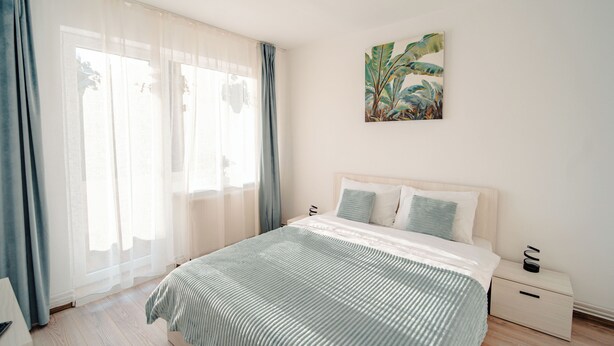 A bedroom with a bed, a nightstand, a wall-mounted painting, and a window with curtains.