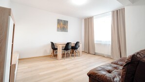A room with a wooden floor, a dining table with chairs, a brown leather sofa, and a large window with curtains.