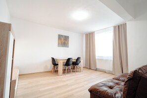 Une pièce avec un plancher en bois, une table à manger avec des chaises, un canapé en cuir marron et une grande fenêtre avec des rideaux.