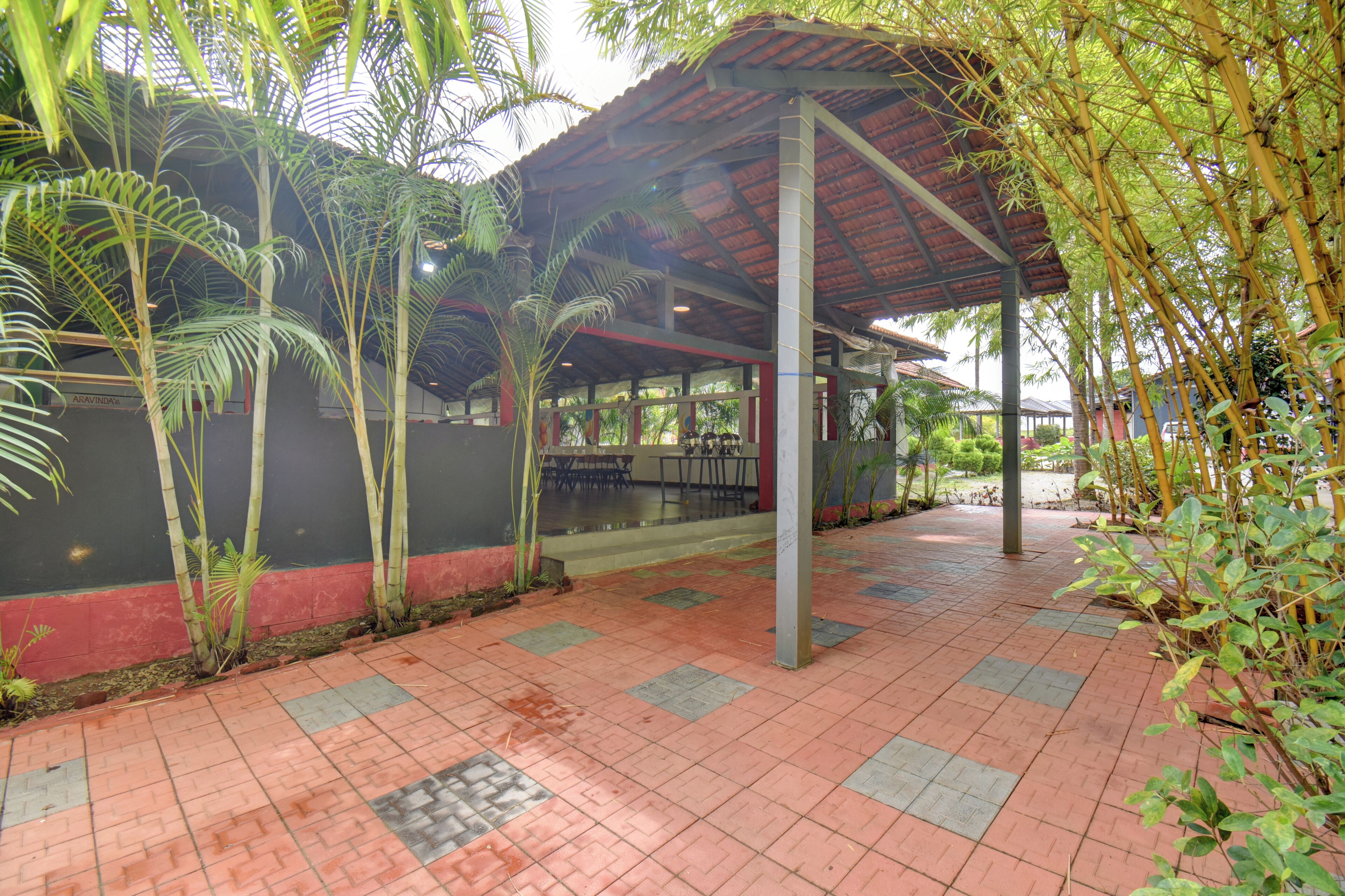 A covered outdoor area with a tiled floor, surrounded by greenery and a building with a red and black facade.