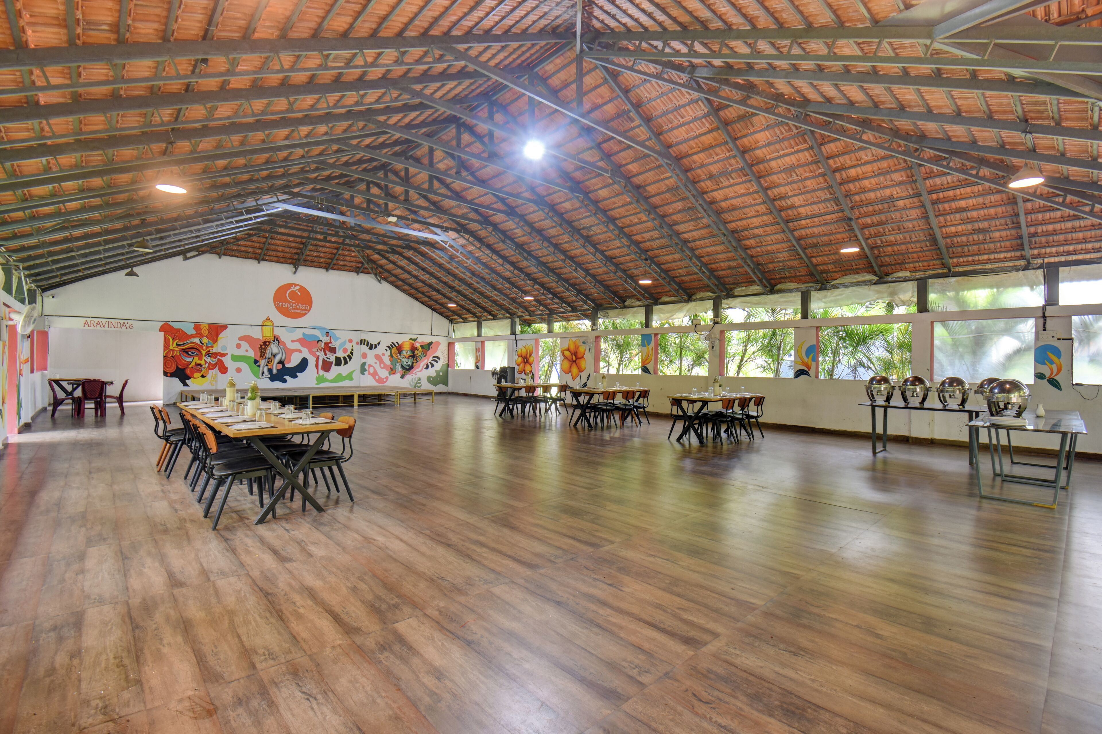 A spacious hall with high ceilings and a wooden floor, featuring tables and chairs arranged for dining.