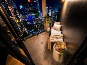 Ein Balkon mit einem Bett, einer Topfpflanze und Blick auf eine nächtliche Stadtlandschaft.