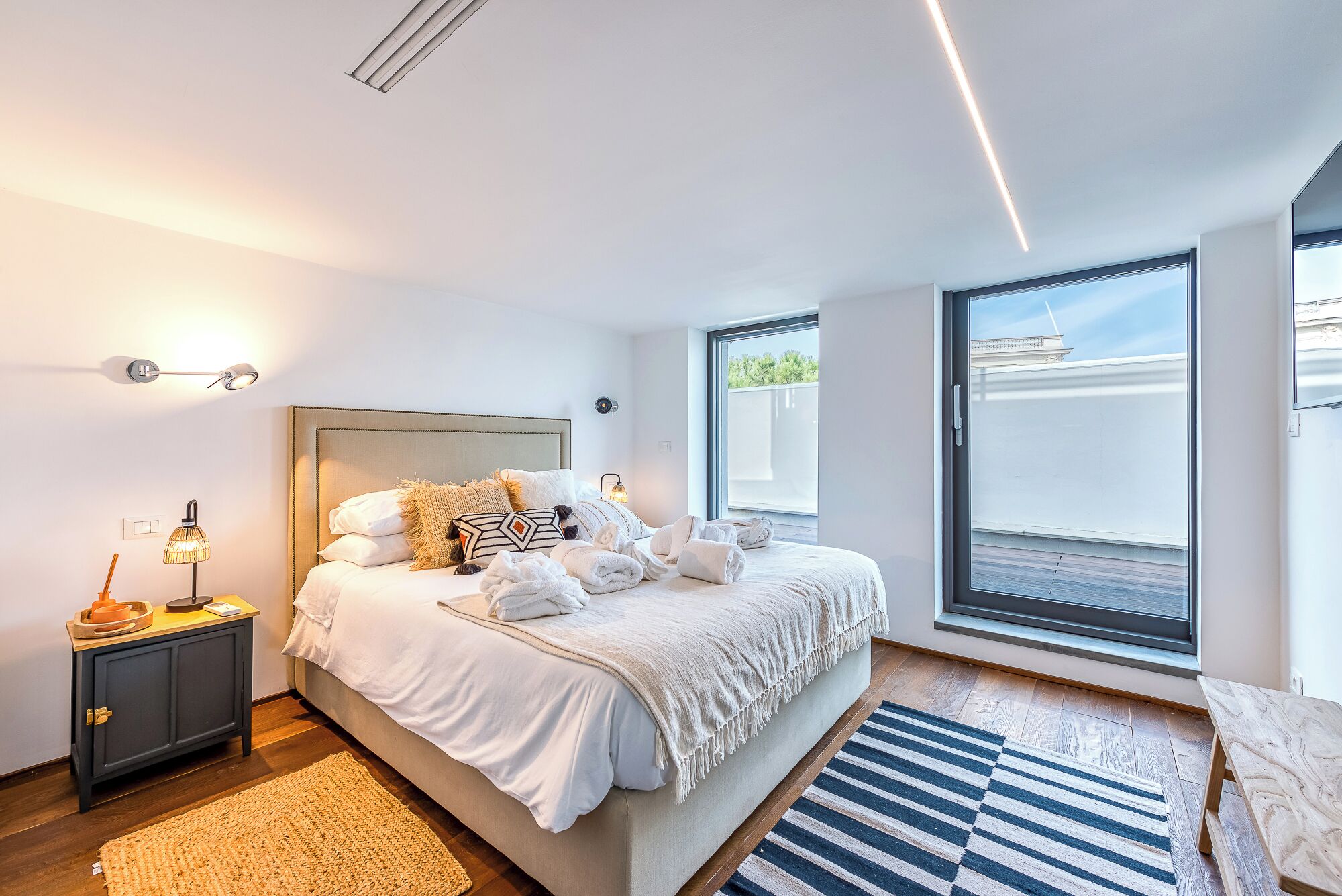 A modern bedroom with a large bed, bedside tables, a nightstand, and a view of the outside.
