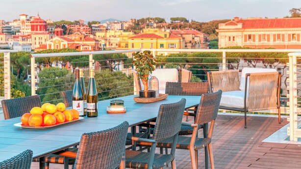 Eine Terrasse mit Esstisch, Korbstühle, einer Weinflasche und einem Blumentopf.