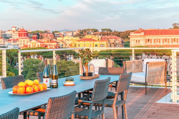 Eine Terrasse mit Esstisch, Korbstühle, einer Weinflasche und einem Blumentopf.